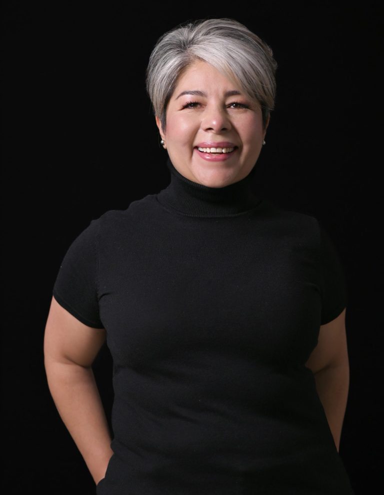 Comunicación Soberanis Mujer sonriente con cabello corto y canoso, vestida con camiseta negra. Fondo oscuro. Es Enid Alvarez Soberanis
